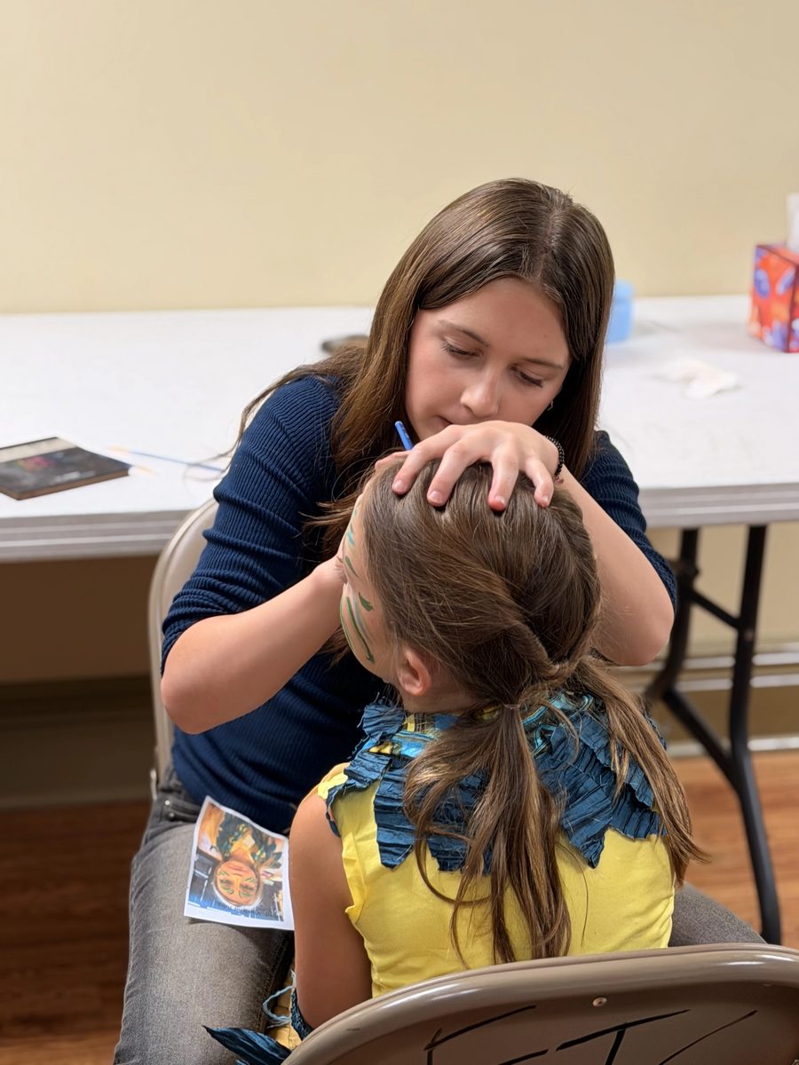 A volunteer paints leaf designs on a young castmate’s face
