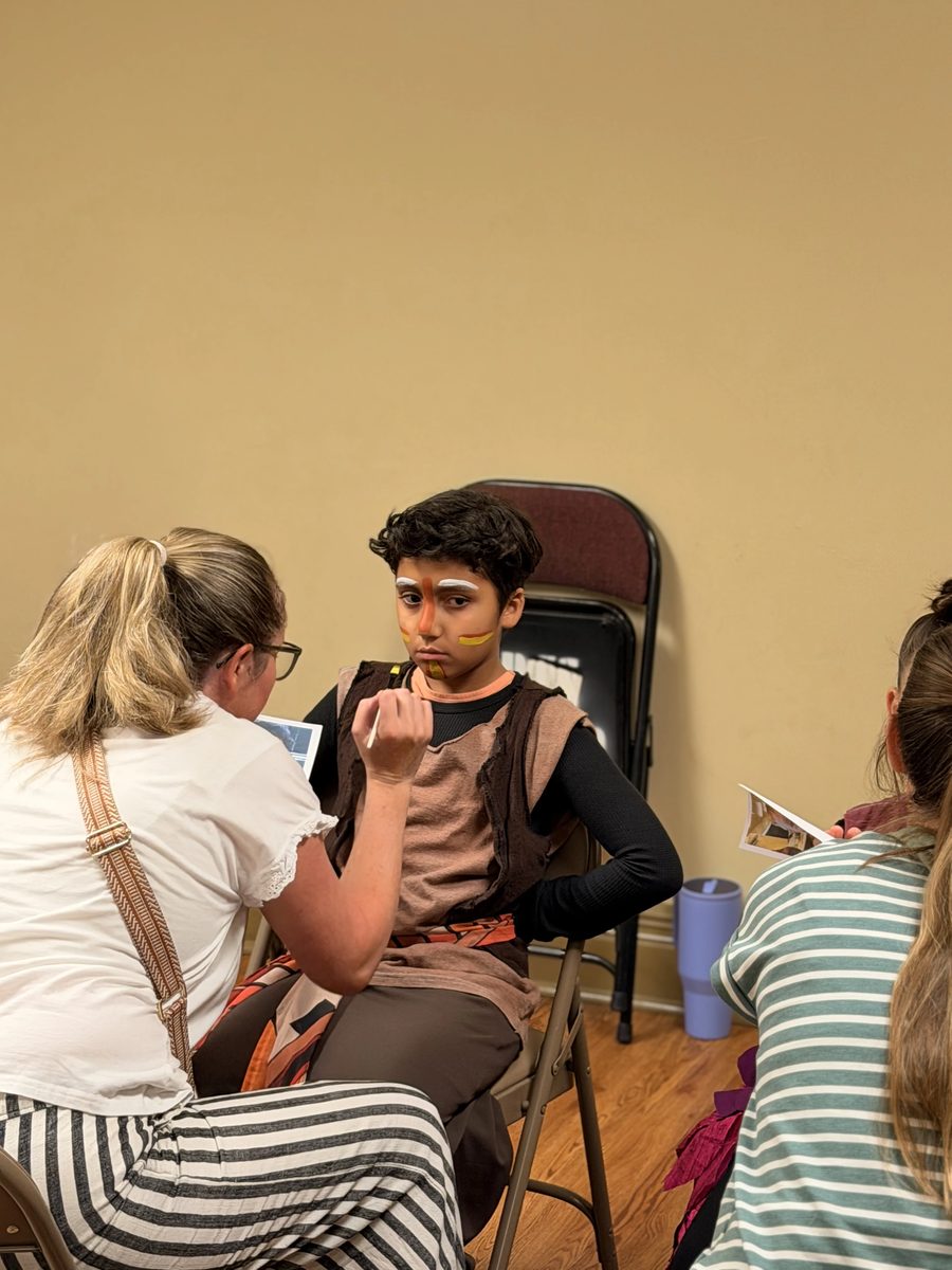 A volunteer applies face paint to a Young Artist in costume