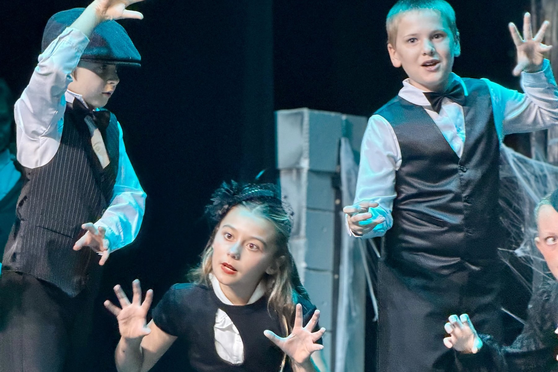 Three Encore Young Artists in black and white costumes reaching toward the audience under blue light during Grim Grinning Ghosts