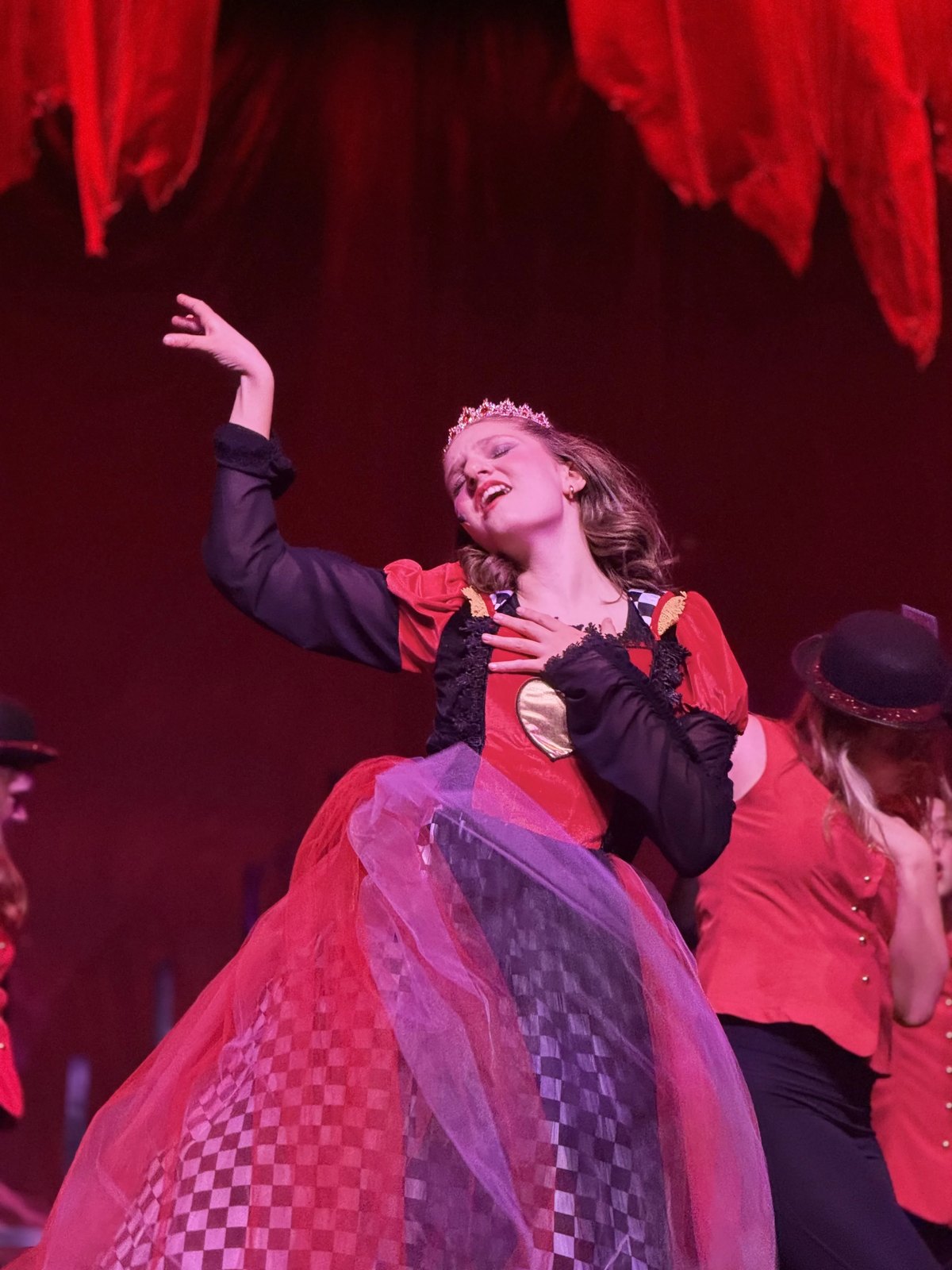 An Encore Young Artist performing as the Queen of Hearts in a red and black gown under red stage light