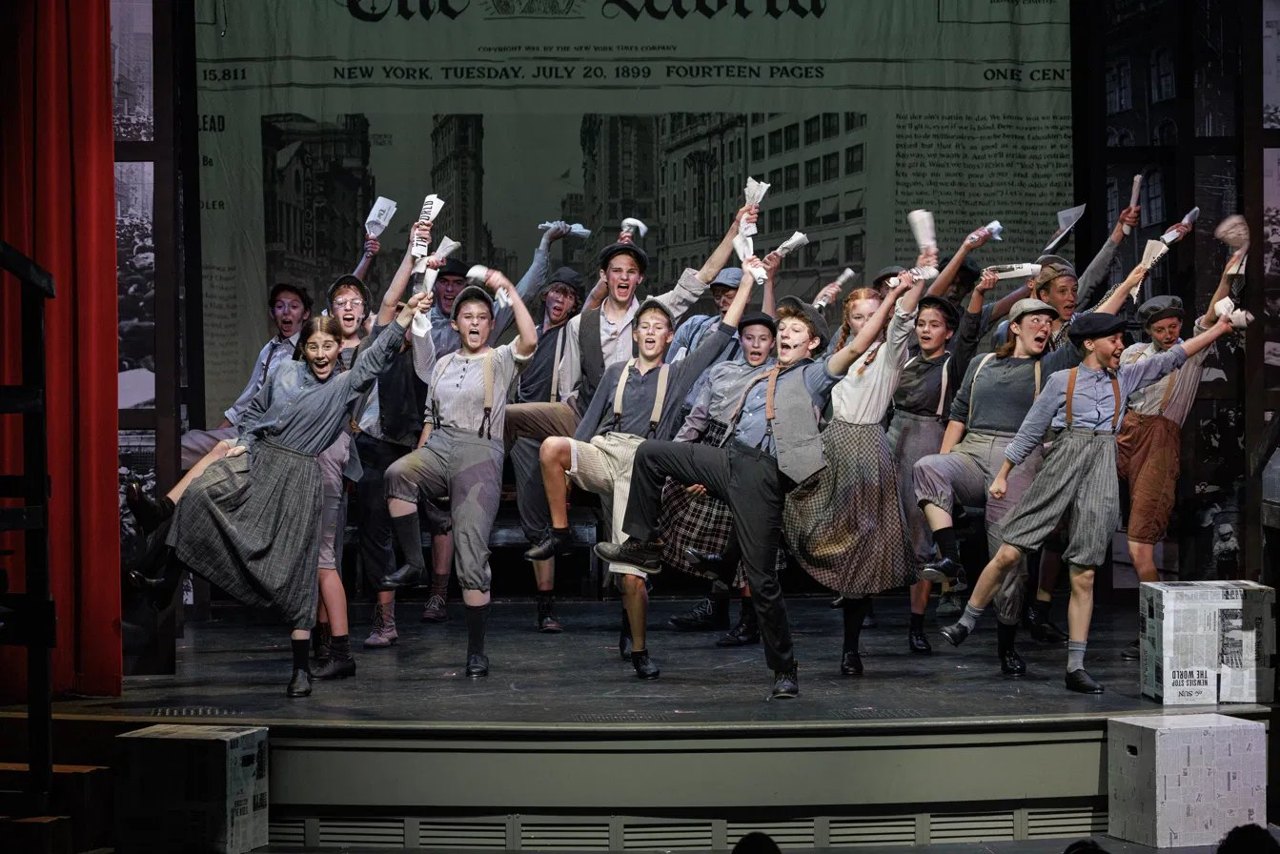 Encore's cast performing a dance number from Newsies in front of a giant newspaper backdrop